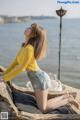 A woman in a yellow sweater and denim shorts sitting on a log by the water.