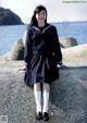 A woman in a school uniform sitting on a rock by the water.